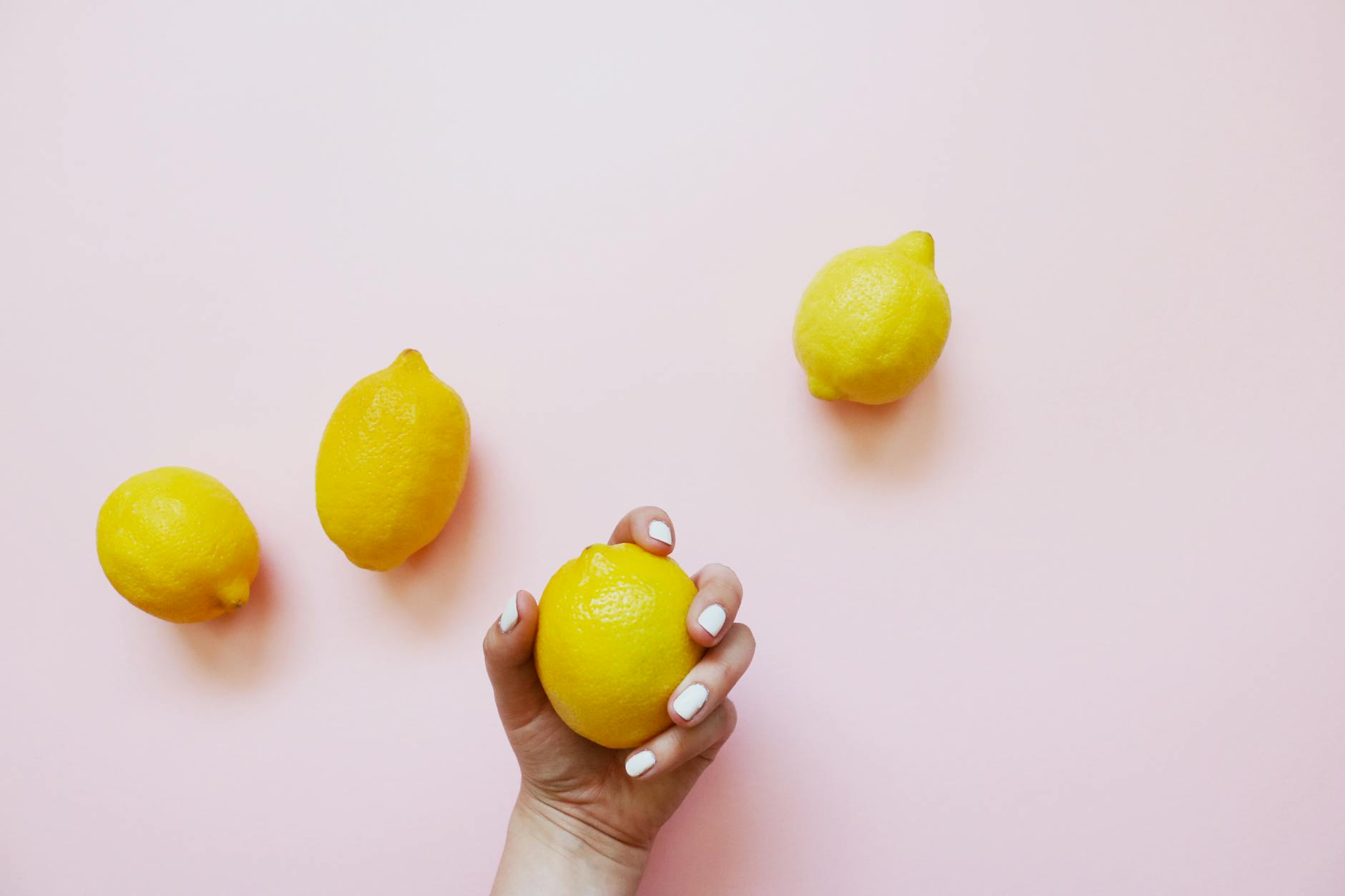 Una mano con smalto bianco che tiene un limone su uno sfondo rosa morbido, circondata da tre limoni aggiuntivi